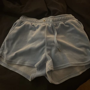 Ljusblå velourshorts från Lindex - Mysiga ljusblå shorts i mjuk velour från Lindex. De har elastisk midja med snörning och sportig look med rundade kanter. Perfekta för chill dagar eller träning. Snyggt och enkelt att matcha med en hoodie eller t-shirt.