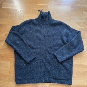 Mörkgrå stickad kofta från Zara - Mysig mörkgrå stickad kofta från Zara med hög krage och lång ärm. Perfekt för kyliga dagar och enkel att matcha med olika outfits. Koftan har en rak, avslappnad passform och är tillverkad i ett mjukt ullblandat material.