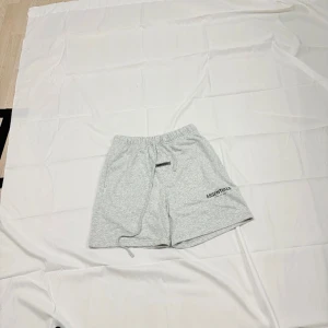 Grå shorts från Essentials Fear of God - Snygga ljusgrå shorts från Essentials Fear of God med elastisk midja och snörning. Mjukt bomullsmaterial, två sidofickor och en bakficka. Diskret logga framtill och patch-detalj. Perfekta för chill eller streetwear.