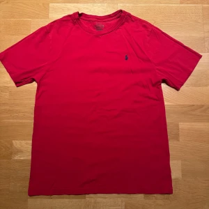 Röd t-shirt från Ralph Lauren - Klassisk röd t-shirt från Ralph Lauren med blå broderad logga på bröstet. T-shirten har rund halsringning och korta ärmar. Tillverkad i mjuk bomull för en skön känsla och enkel stil.
