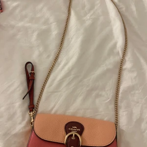 Coach axelväska i rosa och rött skinn - Snygg axelväska från Coach i tvåfärgat skinn, med en mix av ljusrosa och mörkröda toner. Väskan har guldfärgad kedjerem, dekorativt spänne framtill och präglad Coach-logga. Perfekt storlek för det viktigaste och en riktigt trendig accessoar.