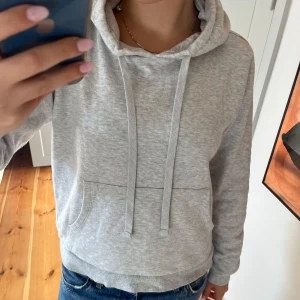 Grå hoodie med huva och ficka - En basic ljusgrå hoodie med huva och dragsnören. Tröjan har en ficka framtill och ribbade muddar vid ärmslut och nederkant. Perfekt för chill dagar eller när du vill ha något bekvämt och enkelt. Står att storleken är L men tycker att den snarare sitter som M