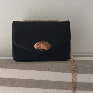 Svart axelväska med guldkedja - Stilren svart axelväska med guldfärgad kedja och snyggt vridlås i guld på framsidan. Väskan har två innerfack varav ett med dragkedja och ett utan. Den är tillverkad i ett läderliknande material som ger en klassisk look. Perfekt storlek för det viktigaste och enkel att matcha till olika outfits. (Aldrig använd)