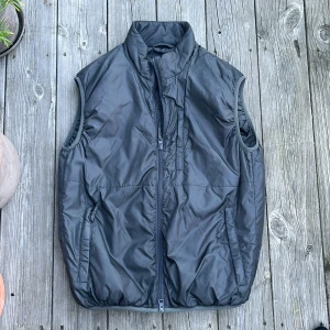 Aspesi väst - Svart dunväst från Aspesi i storlek XS. Västen har hög krage, två sidofickor med dragkedja och en bröstficka med dragkedja. Tillverkad i glansig polyester med quiltad känsla och hel dragkedja framtill.