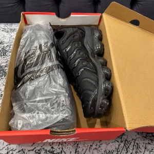 Nike Vapormax svarta sneakers - Nike Vapormax sneakers i helsvart med chunky, transparent sula och snygga detaljer på ovandelen. Skorna har snörning och en modern, sportig look med rund tå och platt sula. Perfekta för dig som gillar streetstyle och vill ha något som sticker ut.