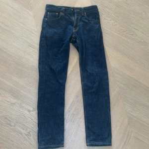 Mörkblå jeans från Nudie Jeans Co 29/32 - Snygga mörkblå jeans från Nudie Jeans Co i klassisk rak modell. Jeansen har kontrastsömmar, fem fickor och Nudie-logga på bakfickan. Tillverkade i slitstark bomull med en stilren look som passar till det mesta.
