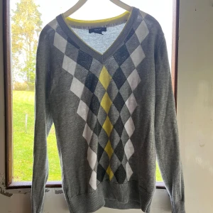 Grå rutig tröja från Abacus - Snygg grå tröja från Abacus med klassiskt rutig mönster i vitt, mörkgrått och gult framtill. V-ringad hals med gul kant och långa ärmar. Perfekt för dig som gillar preppy stil och vill sticka ut lite extra.