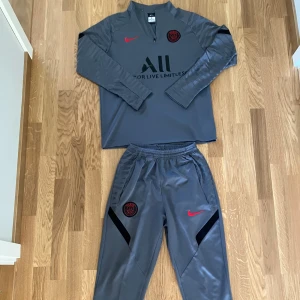 PSG Nike grå set - PSG träningsset från Nike i grått med långärmad tröja och matchande byxor. Tröjan har halv dragkedja, röd Nike-logga och PSG-märke på bröstet. Byxorna har elastisk midja, svarta detaljer och röd Nike-logga. Tillverkad i Dri-FIT material för bästa komfort.
