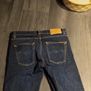 Mörkblå jeans från Nudie Jeans - Snygga mörkblå jeans från Nudie Jeans med klassiska orange sömmar och läderpatch bak. Modellen har raka ben och fem fickor. Jeansen är tillverkade i kraftigt denimtyg av bomull och har en stilren look som passar till det mesta. Pga jeansen är perfekt till dig som är runt 150-160 