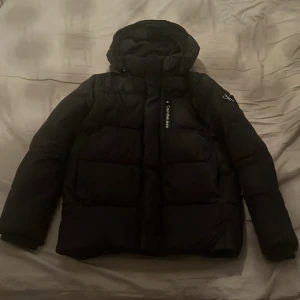 Svart pufferjacka från Calvin Klein Jeans - Svart pufferjacka från Calvin Klein Jeans med huva och logga på ärmen. Jackan har dragkedja framtill med Calvin Klein Jeans-tryck och är vadderad för extra värme. Perfekt för kalla dagar och har en clean, modern look. Skriv gärna vid frågor eller om du vill ha fler bilder!