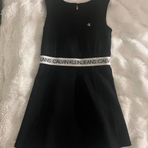 Svart klänning från Calvin Klein - Svart ärmlös kortklänning från Calvin Klein Jeans med vit logotypresår i midjan och liten CK-detalj på bröstet. Klänningen har en enkel, sportig siluett och rund halsringning. Perfekt för dig som gillar stilren och modern design.