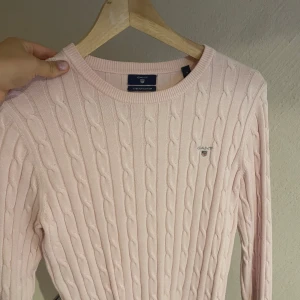 Ljusrosa kabelstickad tröja från Gant - Supersöt ljusrosa kabelstickad tröja från Gant med rund halsringning och broderad logga på bröstet. Tröjan är i stretchig bomull och har långa ärmar. Perfekt för dig som gillar en klassisk och fräsch stil.