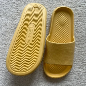 Gula slides sandaler i syntet - Snygga gula slides sandaler med bred rem och öppen tå. Tillverkade i mjukt syntetmaterial med räfflad sula för extra grepp. Perfekta för sommardagar eller att chilla hemma. Enkel och clean design som passar till allt.