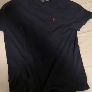 Mörkblå t-shirt från Polo Ralph Lauren - Snygg mörkblå t-shirt från Polo Ralph Lauren med klassisk passform och röd broderad logga på bröstet. Tillverkad i mjuk bomull och har korta ärmar. Perfekt för en clean och stilren look.
