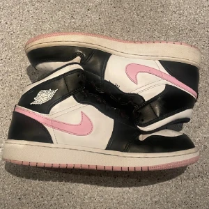 Nike Air Jordan 1 svart/vit/rosa - Säljer ett par Nike Air Jordan 1 med svart och vit bas, rosa swoosh och detaljer. Klassisk high-top siluett med snörning och rund tå. Skorna är i skinn och har rosa yttersula som matchar swooshen. Perfekta för dig som vill sticka ut med en unik färgkombo. Pris kan diskuteras!!🥰