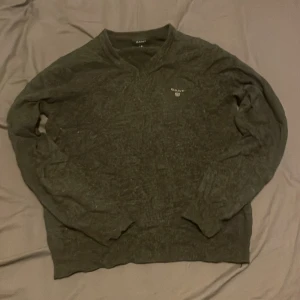 Mörkgrön stickad tröja från GANT - Snygg mörkgrön stickad tröja från GANT med v-ringad hals och diskret logga på bröstet. Tröjan har lång ärm och klassisk passform, perfekt för lager-på-lager under hösten och vintern.