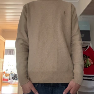 Beige stickad tröja från Polo Ralph Lauren - Snygg beige stickad tröja från Polo Ralph Lauren med diskret broderad logga på bröstet. Tröjan har rund hals, ribbade muddar och är långärmad. Materialet är ull så de är mjukt och skönt mot huden.  Den är som ny så i väldigt bra skick! Hör av er vid frågor eller funderingar :)
