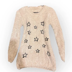 Beige stickad tröja med stjärnor - Mysig beige stickad tröja med svarta stjärnor framtill. Tröjan har lång ärm och en fluffig, lite lurvig känsla i materialet. Perfekt för dig som gillar en chill och trendig look med lite extra detaljer.