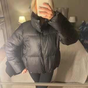 Svart pufferjacka med hög krage - Säljer en svart pufferjacka med hög krage och dragkedja framtill. Perfekt för kalla dagar och har en stilren look som passar till det mesta. Några fläckar innanför kragen men annars i bra skick!