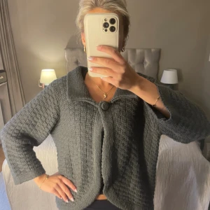 Grå stickad kofta med trekvartsärm - Mysig grå stickad kofta med grov struktur och stor krage. Koftan har en dekorativ knapp framtill och trekvarsärmar.