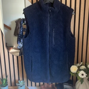 Mörkblå fleeceväst med dragkedja - Mysig mörkblå fleeceväst med hög krage och hel dragkedja framtill. Västen har en bröstficka med dragkedja och två sidofickor. Perfekt för lager-på-lager och chill dagar. Fleece-materialet ger en skön och varm känsla.