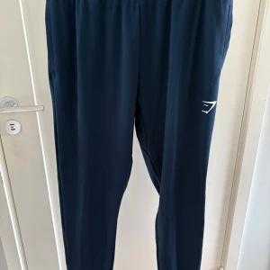 Mörkblå träningsbyxor från Gymshark - Snygga mörkblå byxor från Gymshark med vit logga på benet. Byxorna har elastisk midja och smal passform som sitter perfekt. Mjukt material och diskreta fickor på sidorna.