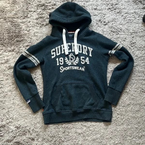 Mörkblå Superdry hoodie med tryck - Mörkblå hoodie från Superdry med vit text och sportiga ränder på ärmarna. Tröjan har huva med vita snören, känguruficka och klassiskt tryck framtill. Riktigt fet, finns lite tecken på användning vilket man märker på trycket. Passar S/M
