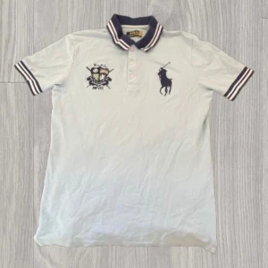 Ljusblå pikétröja från Polo Ralph Lauren - Snygg vit pikétröja från Polo Ralph Lauren med klassisk krage i marinblått och vita ränder. Tröjan har broderad logga och emblem på bröstet, samt korta ärmar med randiga muddar. Perfekt till sommaren och enkel att matcha med jeans eller shorts.