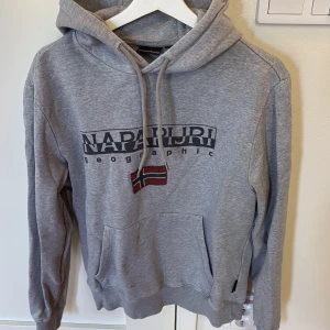 Grå hoodie från Napapijri med tryck - Grå hoodie från Napapijri med stor logga och norsk flagga på bröstet. Tröjan har huva med snörning, magficka och är tillverkad i mjuk bomullsblandning. Perfekt för chill dagar och streetstyle.