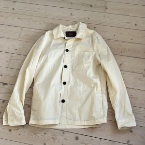 Vit overshirt från Morris, storlek M - Stilren vit overshirt från Morris i bomull. Jackan har svarta knappar framtill och en bröstficka. Perfekt för lager-på-lager och enkel att matcha med det mesta. Den är i storlek M men passar också S. Den har en liten fläck på bröst fickan som borde gå bort i tvätten