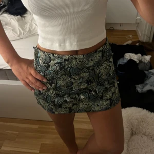 Blommig kortkjol i grönt och svart - Säljer en snygg kortkjol med blommigt mönster i gröna och svarta toner. Kjolen har en tight passform och är perfekt för dig som vill sticka ut med en cool och trendig look. Materialet känns mjukt och följsamt mot kroppen.