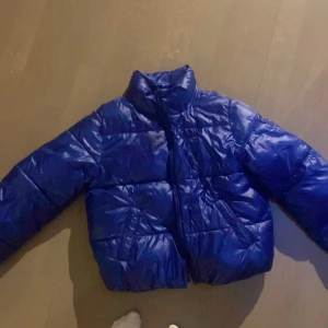 Blå pufferjacka från Tommy Hilfiger - Snygg blå pufferjacka från Tommy Hilfiger med broderad logga på bröstet. Jackan har hög krage, dragkedja framtill och två fickor. Materialet är glansigt och känns riktigt modernt. Perfekt för kalla dagar när du vill hålla stilen. 