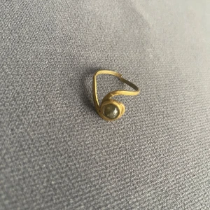 Guldfärgad ring med grå sten - Unik guldfärgad ring med böjd, vågig form och en rund grå sten infattad framtill. Ringen har en öppen design och en modern, konstnärlig känsla som sticker ut. Perfekt för dig som gillar smycken med personlig touch.