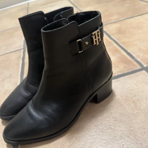 Svarta boots från Tommy Hilfiger - Snygga svarta boots i skinn från Tommy Hilfiger med guldfärgad TH-detalj på sidan. Modellen har spetsig tå, låg klack och dragkedja på insidan. Perfekta för dig som vill ha en stilren och klassisk look med en touch av lyx.