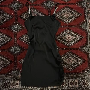Svart kort klänning med smala axelband - En svart, slät och enkel kort klänning med smala axelband. Klänningen har en rak passform och ärmlös design, perfekt för dig som gillar stilren och minimalistisk look. Materialet känns mjukt och följsamt mot kroppen. Super snygga glittriga band och perfekt till halloween+nyår!!