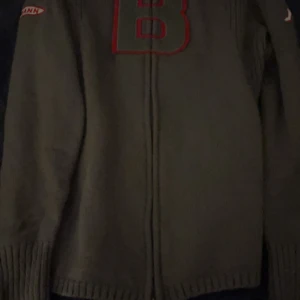 Grå stickad kofta med dragkedja - Säljer en grå stickad kofta från Kappa med dragkedja framtill och stor röd B på bröstet. Tröjan har ribbade muddar och lång ärm, samt broderade detaljer på axeln. Perfekt för dig som gillar avslappnad stil.