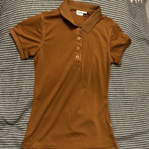 Brun rid t-shirt från CRW XS - Snygg brun pikétröja från CRW i storlek XS. Tröjan har klassisk krage, korta ärmar och knappar framtill. Perfekt för dig som gillar en stilren och enkel look. Materialet känns mjukt och följsamt mot kroppen.