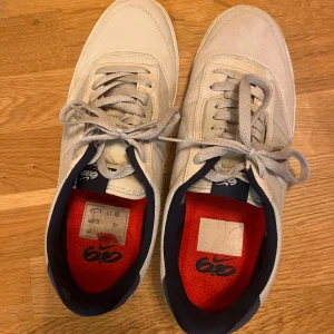 Nike vita sneakers med marinblå detaljer - Snygga vita Nike sneakers med marinblå detaljer och röd innersula. Skorna har klassisk snörning, rund tå och platt sula. Ovandelen är i textil och mocka för en stilren look. 