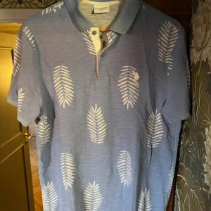 Blå piké med vita blad från Polo Ralph Lauren - Snygg blå pikétröja från Polo Ralph Lauren med vita bladmönster över hela tröjan. Klassisk krage och korta ärmar, perfekt för en avslappnad stil. Skön bomullskvalitet och diskret logga på bröstet.