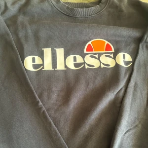 Mörkgrå sweatshirt från Ellesse - Säljer en mörkgrå sweatshirt från Ellesse med stort vitt logotryck och orange detalj framtill. Tröjan har rund halsringning och långa ärmar, perfekt för en chill och sportig look. Materialet är mjukt och skönt, troligtvis bomullsmix.