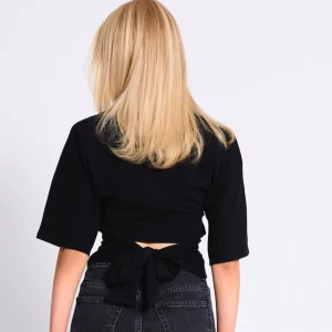 Svart topp med knyt och kort ärm - Svart topp i bomull med korta ärmar och rund hals. Toppen har en snygg knytning i midjan som ger en cool detalj baktill. Passformen är normal och materialet känns mjukt mot huden. Perfekt till jeans eller kjol för en stilren look.använd bara en gång 