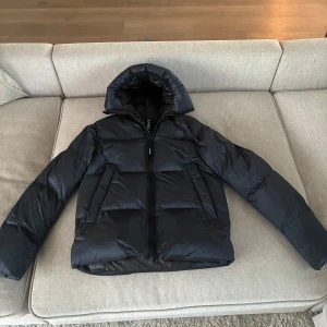 Mörkblå pufferjacka från Canada Goose - Säljer en Mörkblå pufferjacka från Canada Goose med huva och dragkedja. Jackan har två fickor framtill och reflekterande detaljer på huvan. Perfekt för kalla dagar och riktigt snygg streetstil. Fodrad med dun för extra värme. Kom med frågor om ni är intresserade! Pris kan diskuteras!