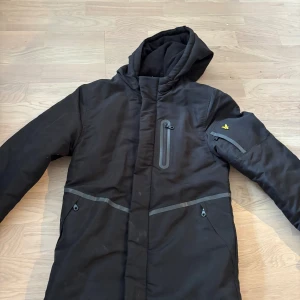 Svart vinterjacka från Lyle & Scott - Säljer en svart vinterjacka från Lyle & Scott med huva och fleecefoder. Jackan har dragkedjor på fickorna, bröstficka och en diskret logga på ärmen. Perfekt för kalla dagar och har en clean look med reflexdetaljer.