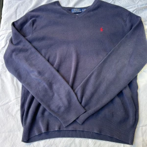 Blå v-ringad tröja från Polo Ralph Lauren - Snygg marinblå v-ringad tröja från Polo Ralph Lauren i mjuk pima bomull. Klassisk design med röd broderad logga på bröstet och ribbade muddar vid ärmslut och nederkant. Perfekt för dig som gillar stilrena och tidlösa plagg.