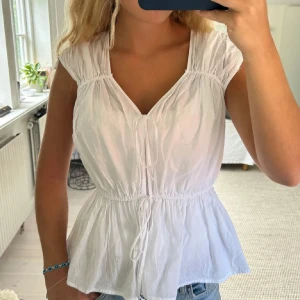 Vit blus med knyt och v-ringning - Söt vit blus med v-ringning och knyt framtill. Modellen är ärmlös med lätt rynkade axlar och markerad midja med knytband. Luftigt bomullstyg som passar perfekt till jeans eller kjol. Enkel och fräsch stil för alla tillfällen.