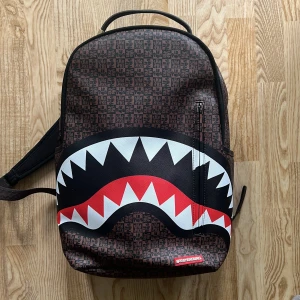 Sprayground ryggsäck med hajmotiv - Cool ryggsäck från Sprayground med hajtandsmönster framtill och brunt rutigt mönster över hela väskan. Ryggsäcken har rödfärgat innerfoder, flera fack och justerbara axelremmar. Perfekt för dig som vill sticka ut med en unik och trendig accessoar. Kvitto finns, köpt från italien