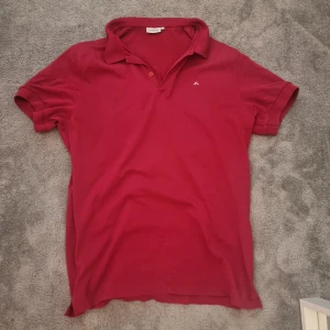 Röd pikétröja från J.Lindeberg - Röd pikétröja från J.Lindeberg med klassisk krage, kort ärm och liten vit logga på bröstet. Lite slitage vid kragen och vänstra armhåla men är knappast synliga.