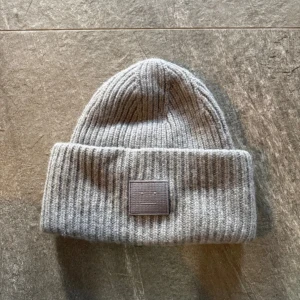 Grå ribbstickad mössa Acne Studios - Säljer en grå ribbstickad mössa från Acne Studios med klassisk uppvikt kant och den ikoniska Acne Studios-loggan framtill. Mössan är tillverkad i mjukt ullmaterial och har en enkel, stilren design som passar perfekt till kalla dagar.