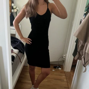 Svart tight klänning med smala axelband - En svart, tight klänning med smala axelband och v-ringning. Klänningen är kort och har en enkel, stilren design som sitter figurnära. Perfekt för dig som gillar minimalistisk stil och vill ha något som framhäver siluetten.