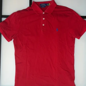 Röd pikétröja från Ralph Lauren - Klassisk röd pikétröja från Ralph Lauren med blå broderad logga på bröstet. Tröjan har krage, tre vita knappar och korta ärmar. Tillverkad i mjuk bomull som är skön mot huden. Perfekt för en stilren och sportig look.